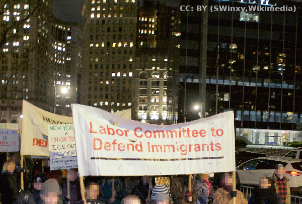 abendliche Demo mit Banner: Labor Committee to Defend Immigrants; im Hintergrund beleuchtete Gebäude in New York City